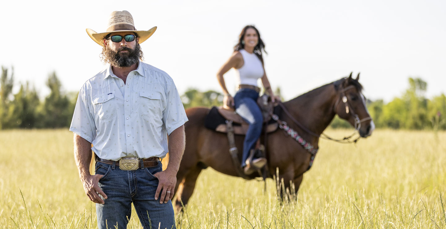 Kip Smith with a cowboy hat and beard, standing in front in a field, while Kalie Smith is riding a brown horse in the background in the field. 
